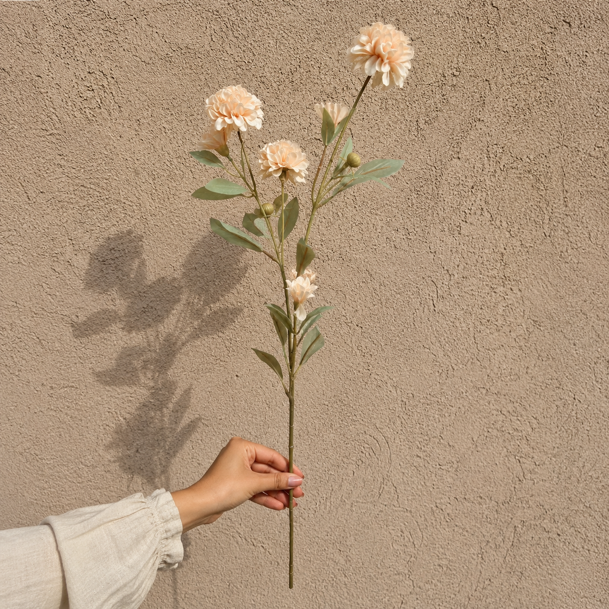 Woman’s hand holding a single artificial peach carnation stem against a textured beige wall in natural daylight, showcasing the full stem and flower details.