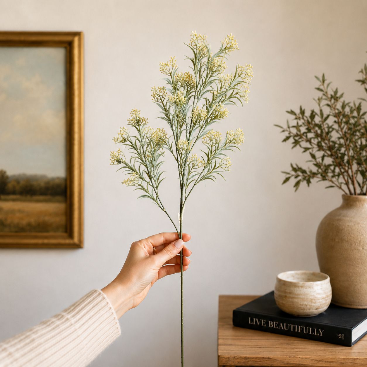 Single artificial green wildflower stem held in hand in a modern home setting, showcasing realistic foliage and delicate blooms against a neutral interior background.