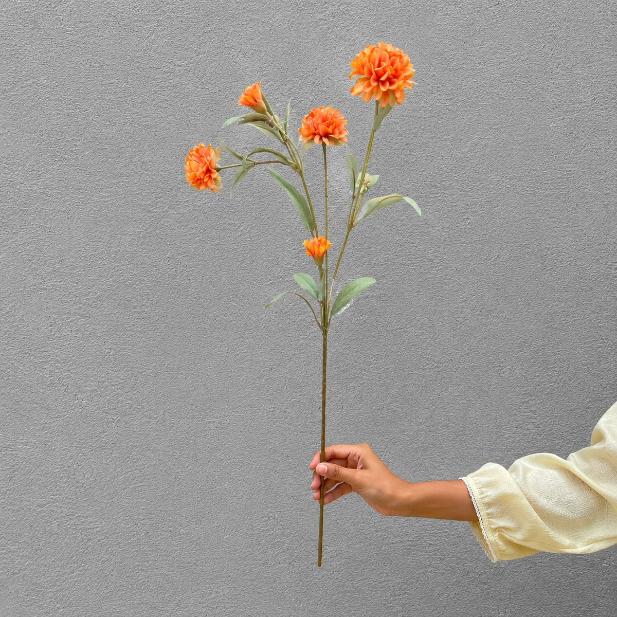Hand holding a single artificial orange carnation stem with multiple blooms against a textured grey wall, showcasing the flower's size and detailing in natural light.
