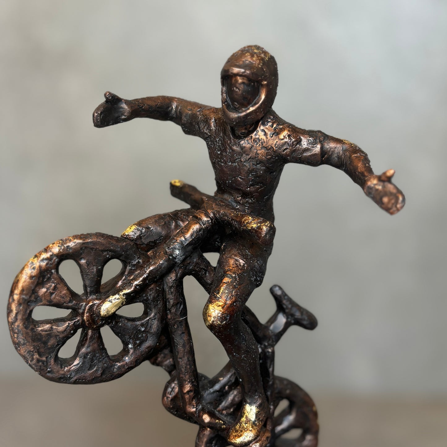 Detailed view of a handcrafted stunt biker sculpture showing the aged bronze texture, helmet detail, and gold-leaf accents on the rider’s joints and bike.