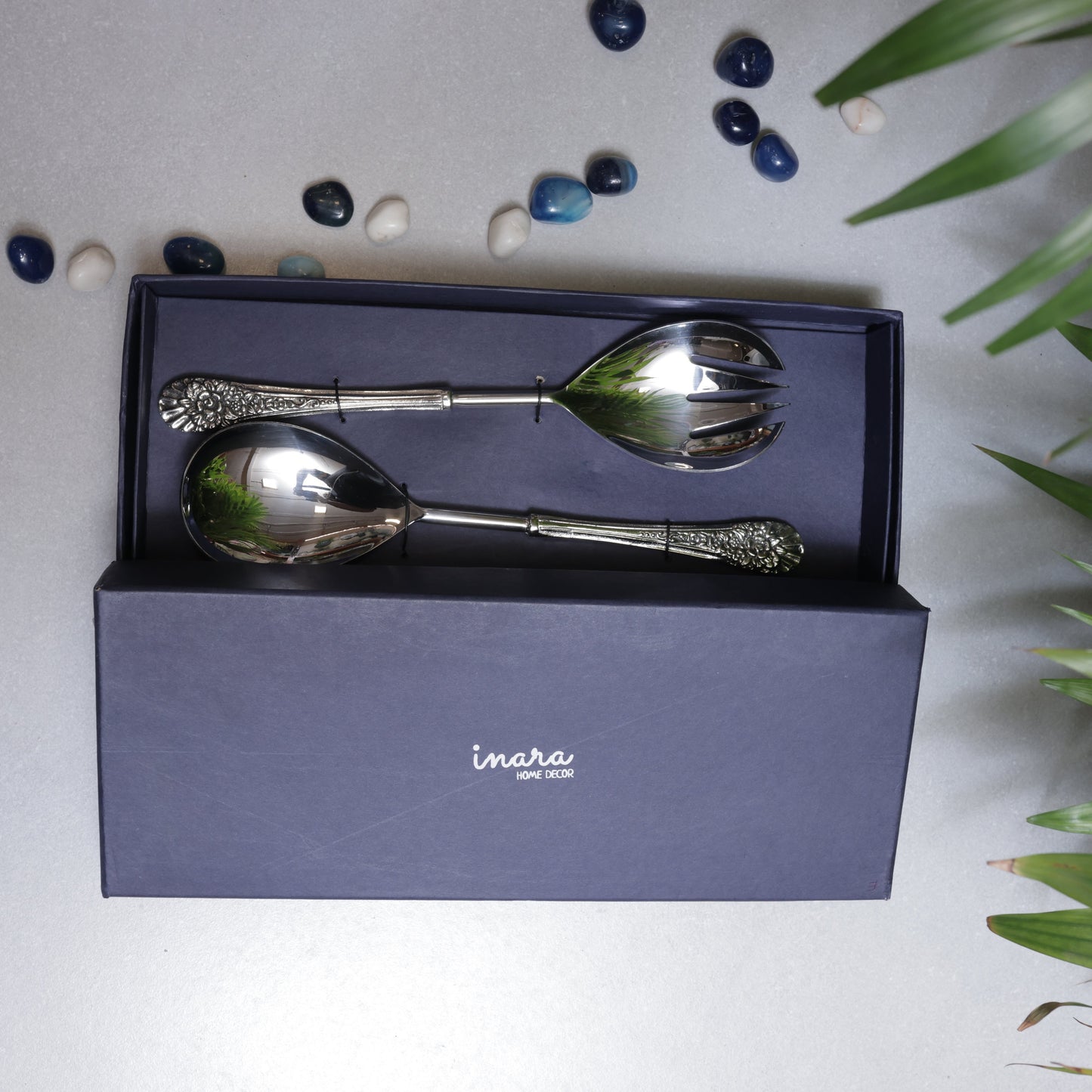 A pair of silver Inara Home Decor salad servers, including a large spoon and a spork, presented in a sleek navy blue branded gift box.