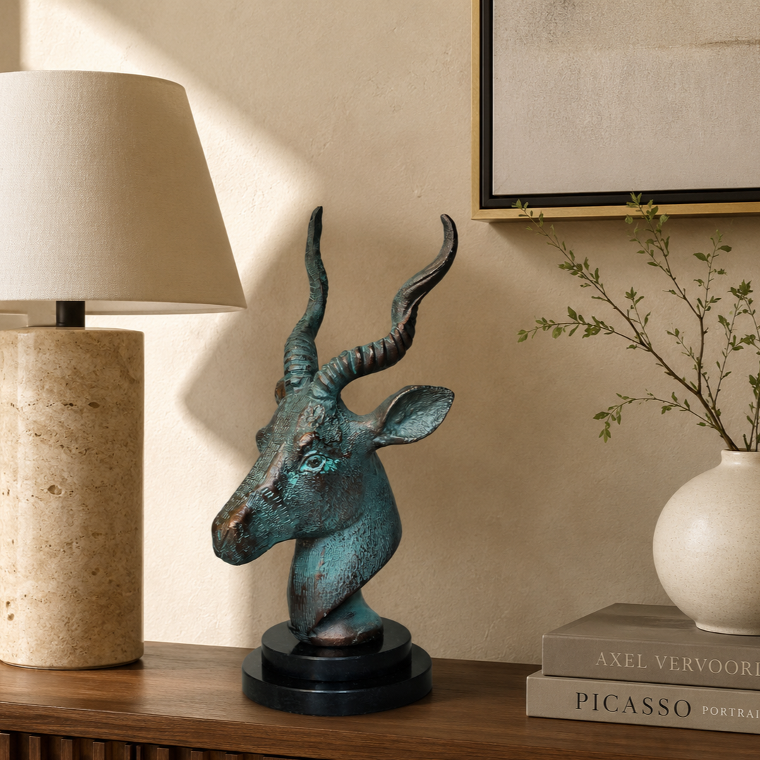 Luxury antelope head sculpture with verdigris finish displayed on a walnut entryway console with a travertine table lamp, ceramic vase, coffee table books, and botanical accents against a warm beige Venetian plaster wall.
