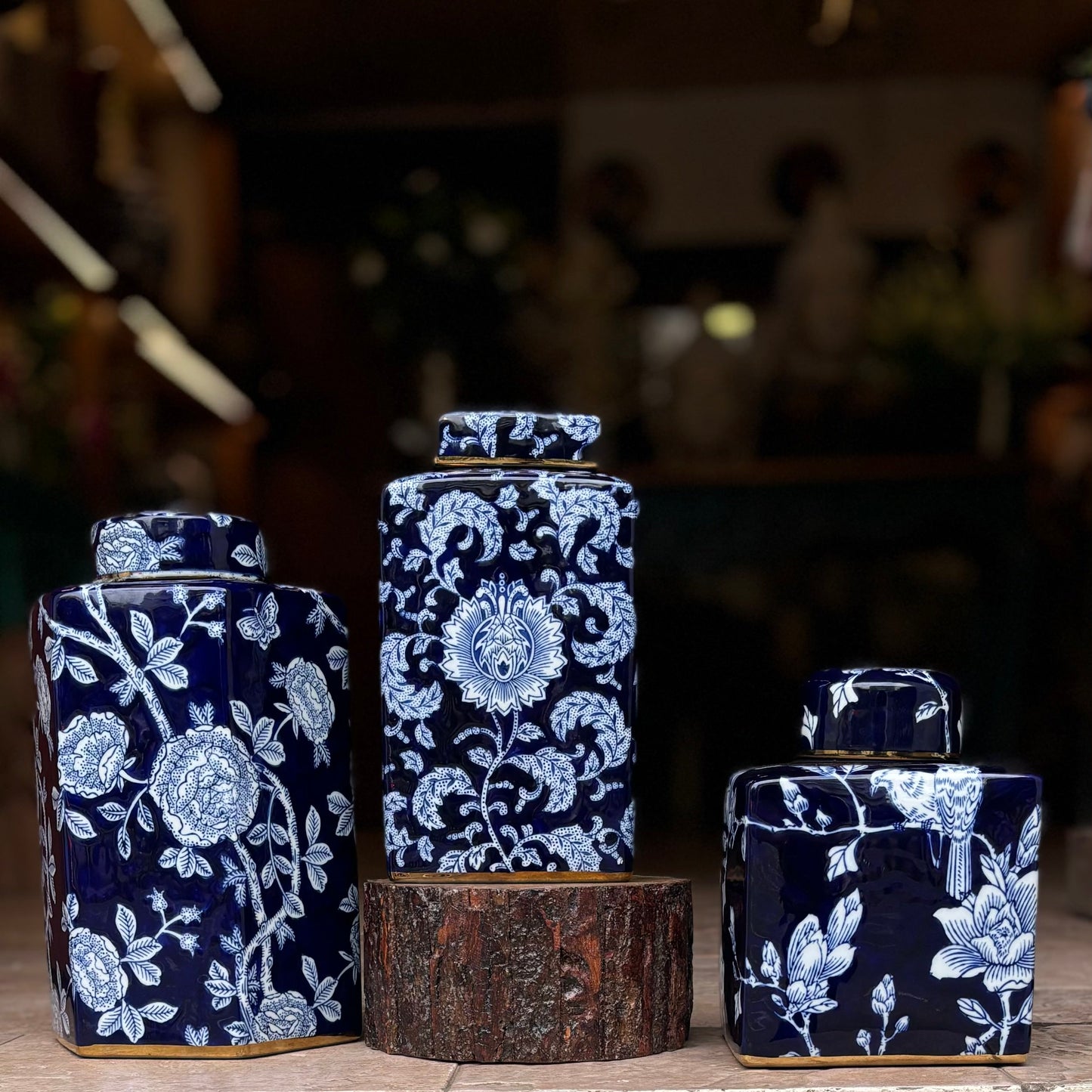 A set of three graduated navy blue and white porcelain ginger jars with intricate floral and botanical patterns, featuring elegant gold-rimmed bases and lids, displayed on a rustic wood pedestal.