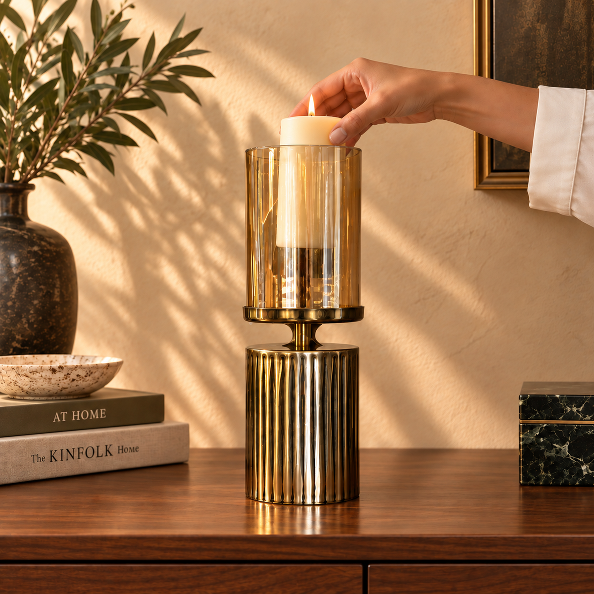 Hand placing a lit cream pillar candle into an amber glass hurricane candle holder with a ribbed gold metal base, demonstrating easy setup and elegant home décor styling.