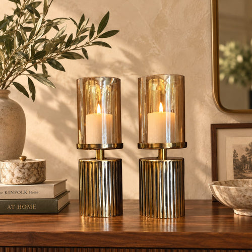 Luxury amber glass hurricane candle holder set with lit pillar candles styled on a walnut console table, featuring a ribbed gold metal base for elegant home décor.