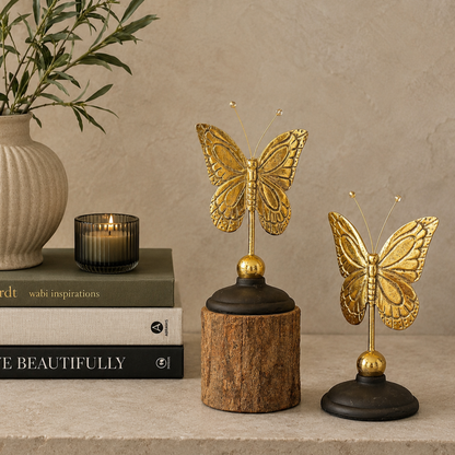 Antique brass butterfly figurines displayed beside stacked luxury coffee table books, a ceramic vase, and a decorative candle on a warm beige backdrop, showcasing their size and elegant home décor styling.