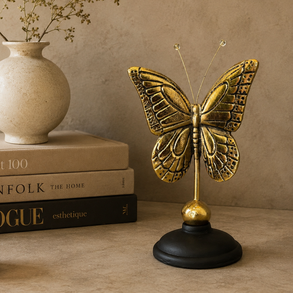 Antique brass butterfly sculpture on a black base displayed beside stacked coffee table books, a ceramic vase, and a decorative candle, showcasing its size in a luxury home décor setting.