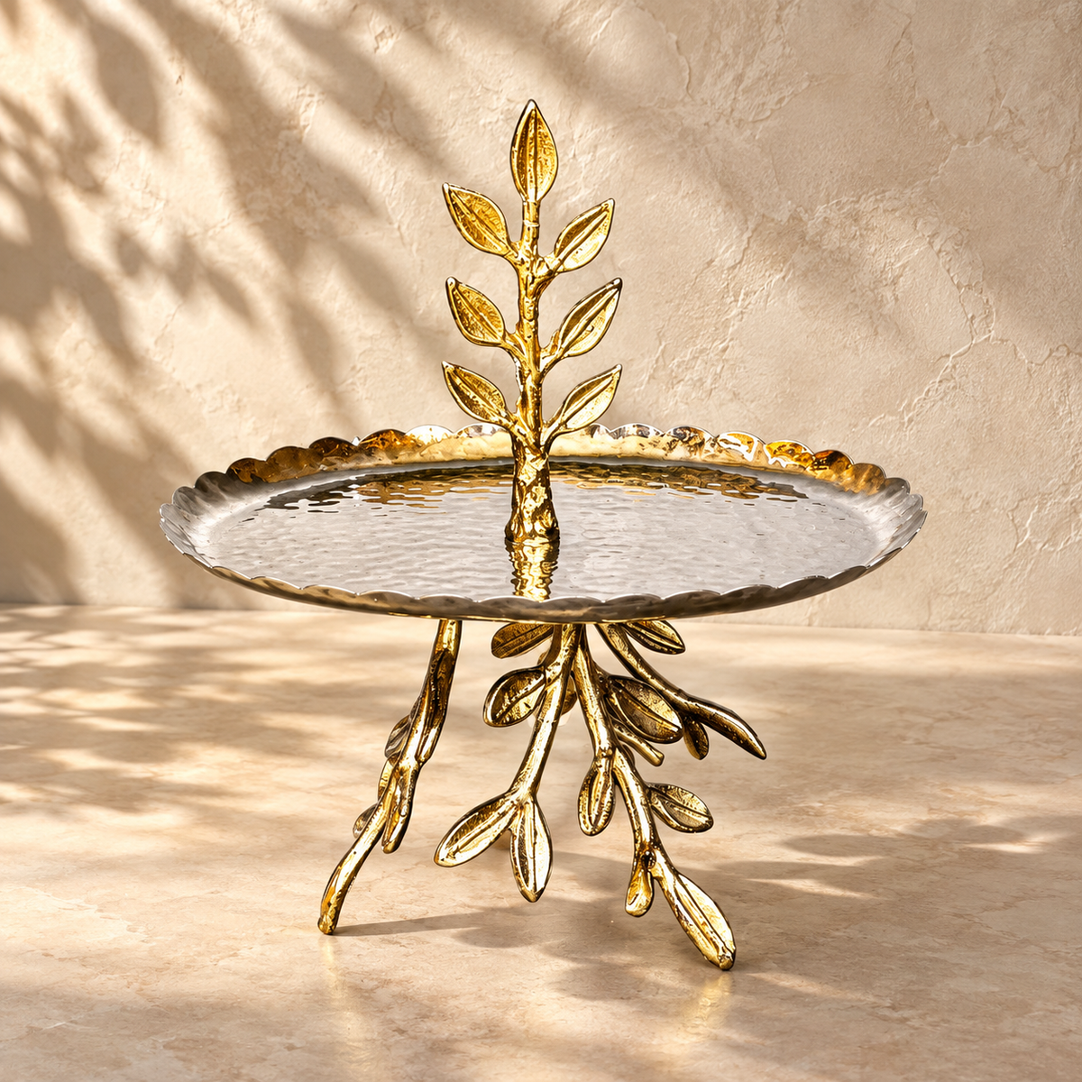 Antique gold leaf pedestal decorative metal serving tray with a hammered scalloped tray, styled as a luxury hero image on a warm beige microcement surface with a Venetian plaster wall and soft natural sunlight.