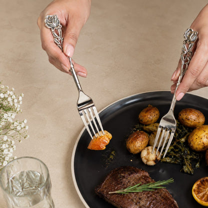 Antique Rose floral stainless steel dinner forks in use during an elegant dining experience on a black plate with gourmet food, styled on a warm beige Venetian seamless matte microcement surface.