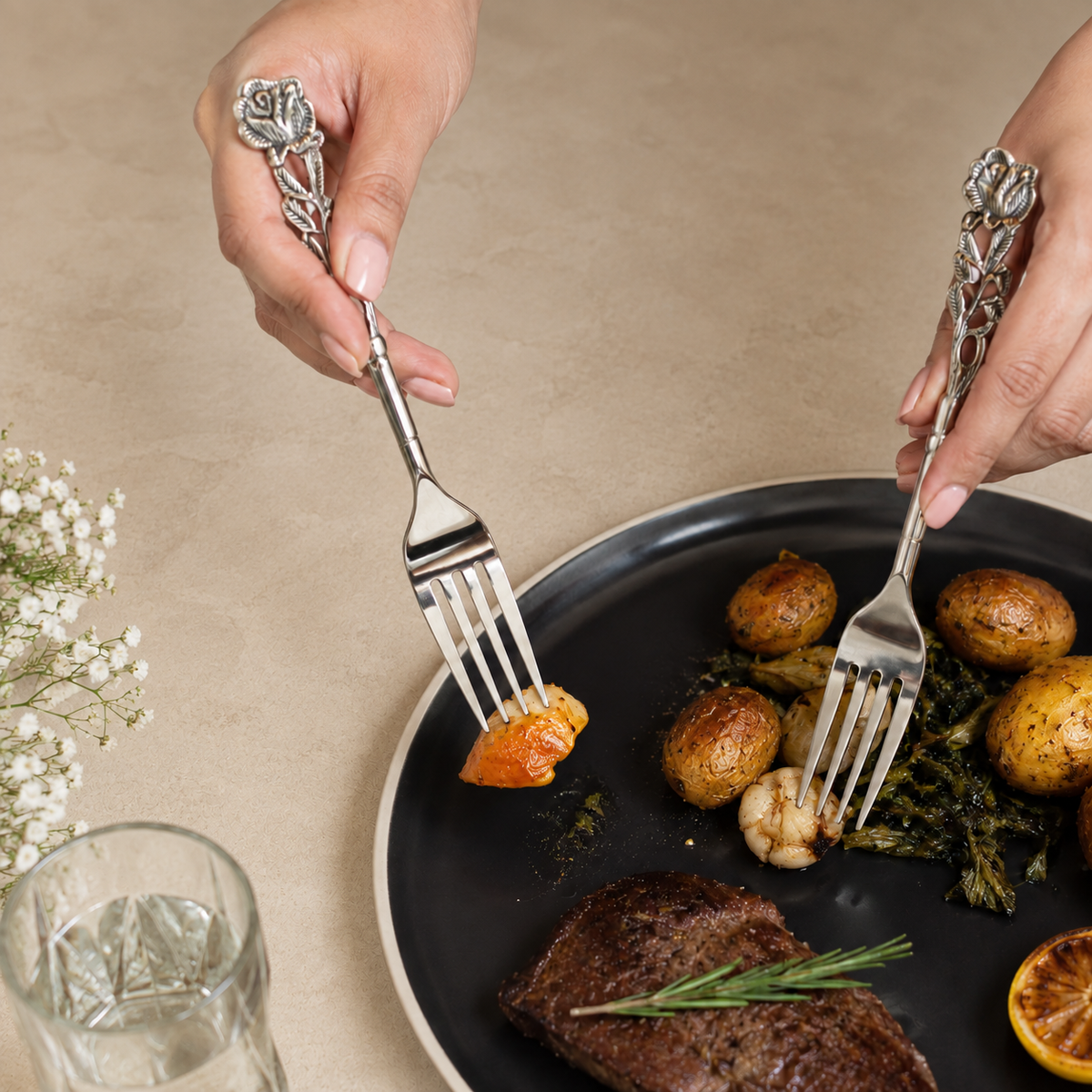 Antique Rose floral stainless steel dinner forks in use during an elegant dining experience on a black plate with gourmet food, styled on a warm beige Venetian seamless matte microcement surface.