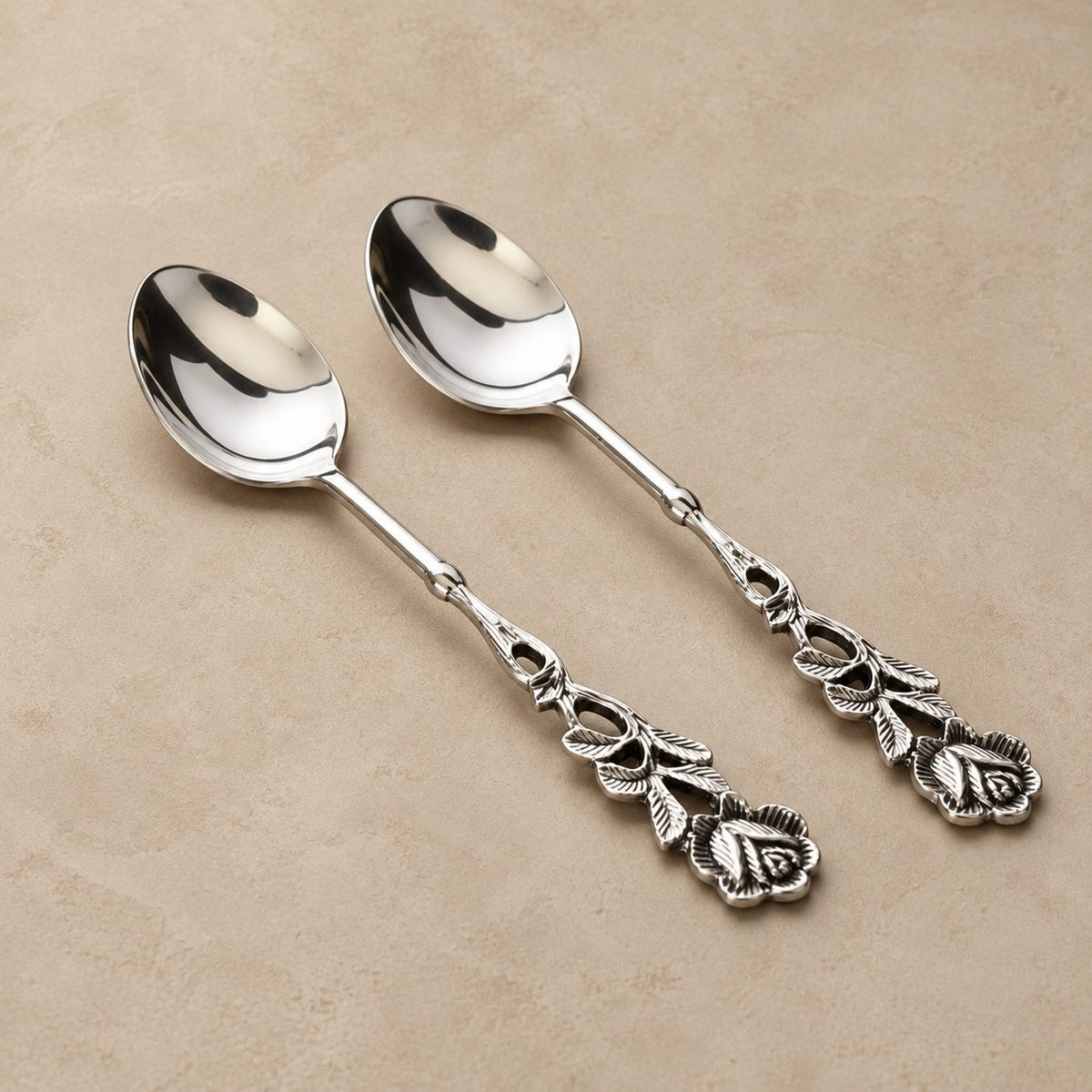 Antique Rose silver dessert spoons set of 2 with ornate floral handles in polished stainless steel, displayed on a warm beige seamless matte microcement surface for luxury table setting.