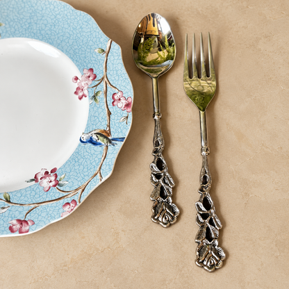 Antique Rose spoon and fork set with ornate floral handles styled beside a blue floral porcelain plate on a warm beige seamless matte microcement surface, luxury flatware by Inara Home Decor.