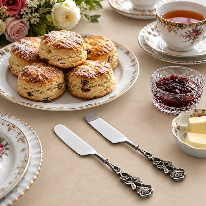 Antique Rose stainless steel butter spreader knives with decorative floral handles displayed on a warm beige Venetian seamless matte microcement surface alongside fresh scones, tea, jam, and fine floral china for an elegant afternoon tea setting.