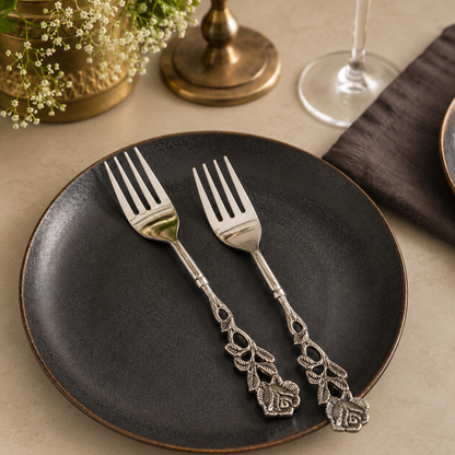 Antique Rose stainless steel dessert forks set of 2 with decorative floral handles displayed on a black ceramic plate over a warm beige Venetian seamless matte microcement tabletop, luxury cutlery by Inara Home Decor.