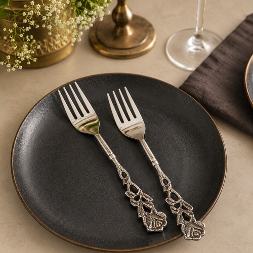Antique Rose stainless steel dessert forks set of 2 with decorative floral handles displayed on a black ceramic plate over a warm beige Venetian seamless matte microcement tabletop, luxury cutlery by Inara Home Decor.