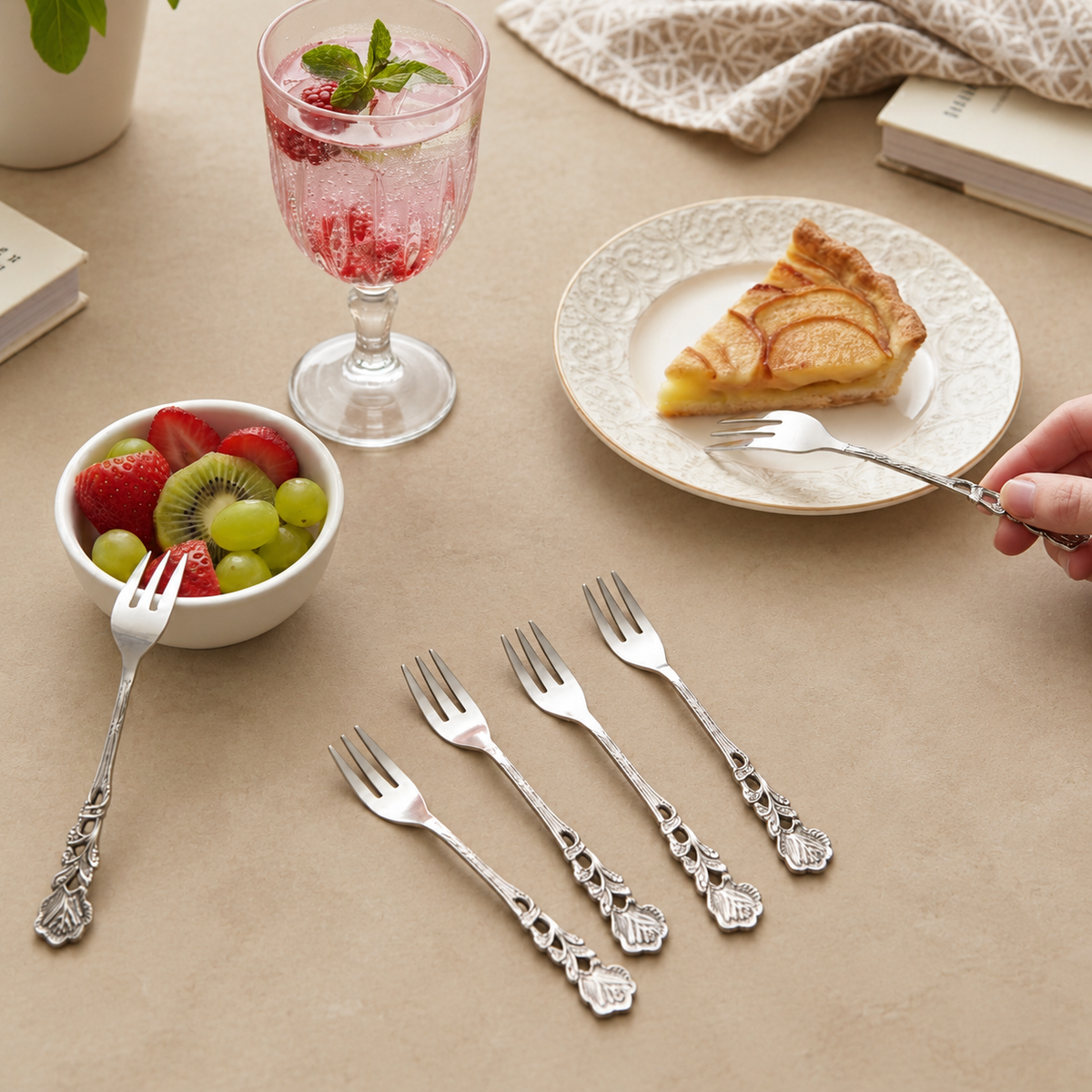 Antique Rose stainless steel dessert forks set of 6 with decorative floral handles, elegantly styled on a warm beige Venetian seamless matte microcement tabletop alongside fresh fruit, sparkling drink, and dessert for a premium dining setting.