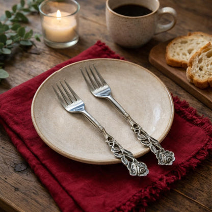 Pair of floral-themed dessert forks placed on a neutral beige ceramic plate and red linen napkin, featuring a rustic coffee and toast setting.