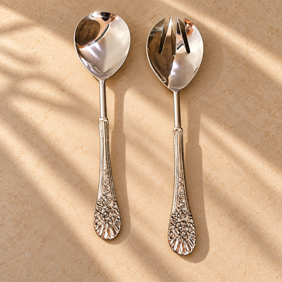 Antique silver floral salad serving spoon and fork set with ornate embossed handles, displayed on a warm beige stone surface in a luxury hero lifestyle setting.