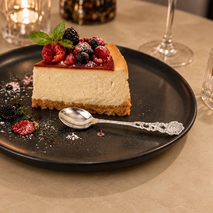 Antique silver rose dessert spoon beside a berry cheesecake on a black plate, styled on a warm beige Venetian seamless matte microcement table in an elegant luxury dining setting.