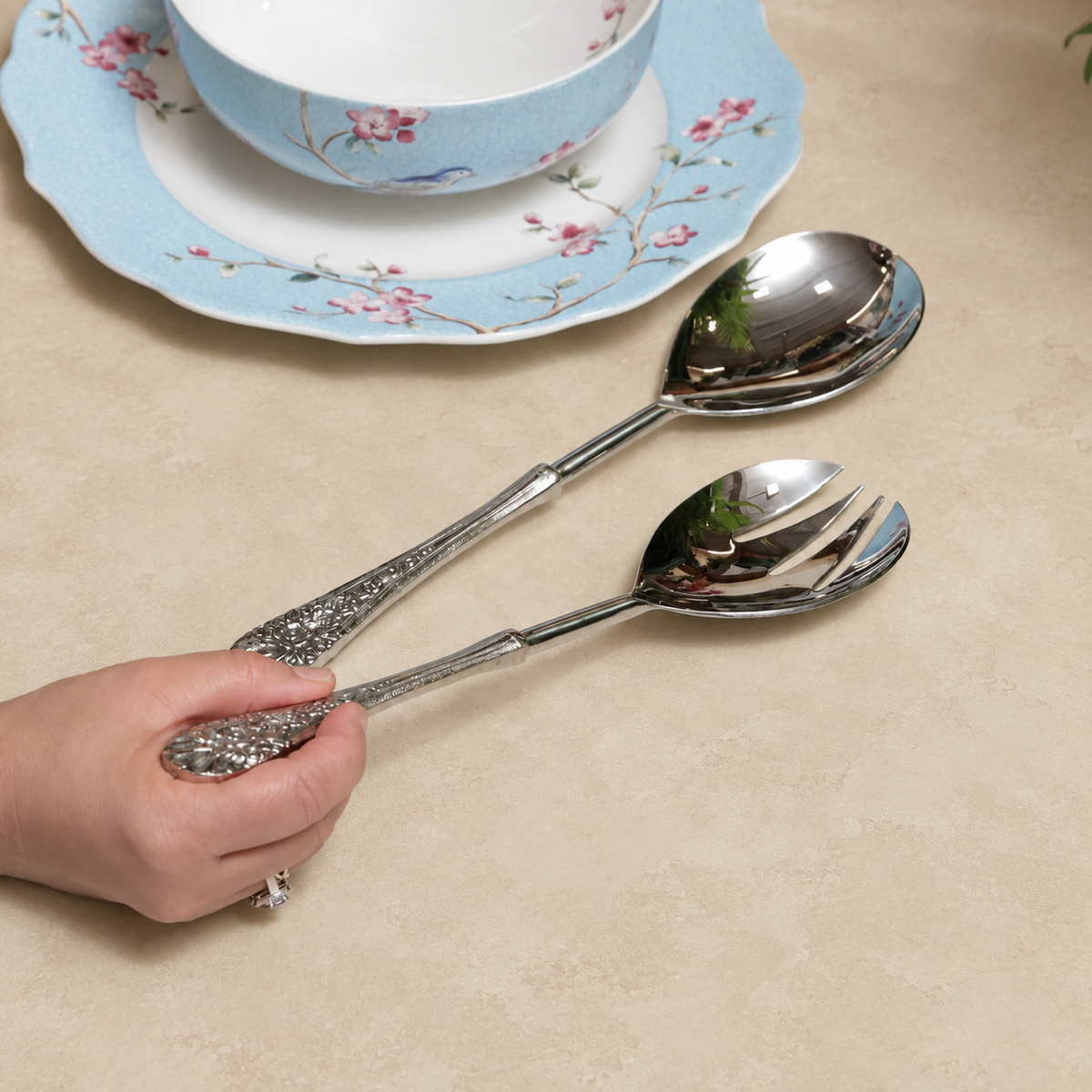 Antique silver serving spoon and fork set with ornate floral handles displayed on a warm beige Venetian seamless matte microcement surface beside elegant blue floral porcelain dinnerware, perfect for luxury dining and serving.