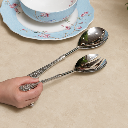 Antique silver serving spoon and fork set with ornate floral handles displayed on a warm beige Venetian seamless matte microcement surface beside elegant blue floral porcelain dinnerware, perfect for luxury dining and serving.