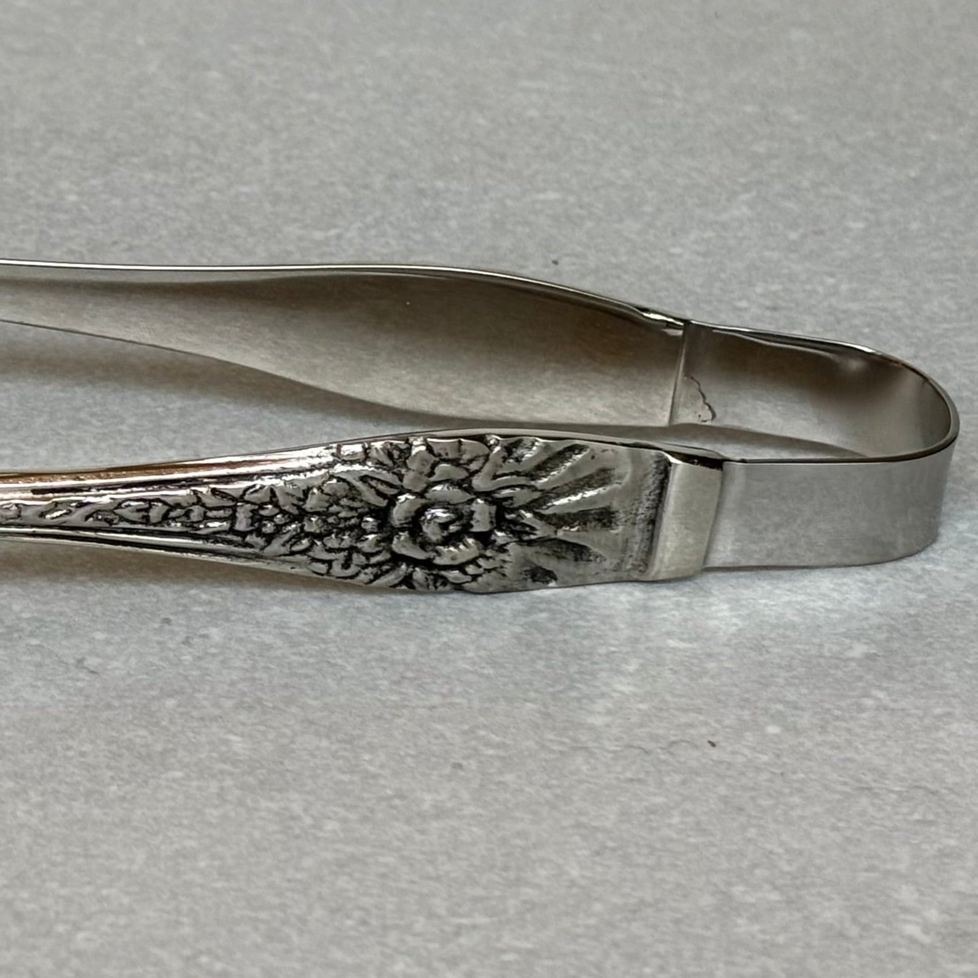 Antique silver serving tongs with intricate floral embossed handle detail and stainless steel body on textured background