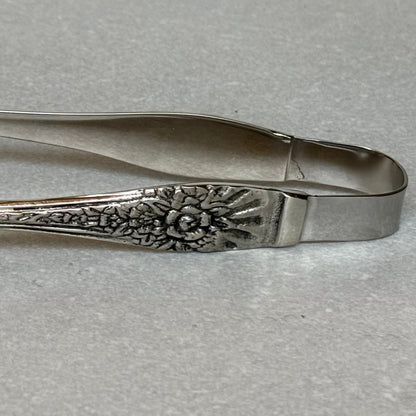 Antique silver serving tongs with intricate floral embossed handle detail and stainless steel body on textured background