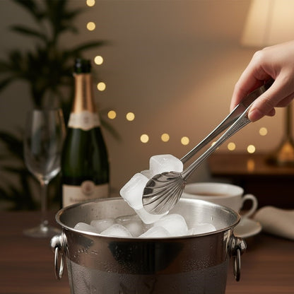 Hand holding antique silver serving tongs lifting ice cubes from a metal ice bucket in an elegant bar setup with champagne bottle and glasses in background