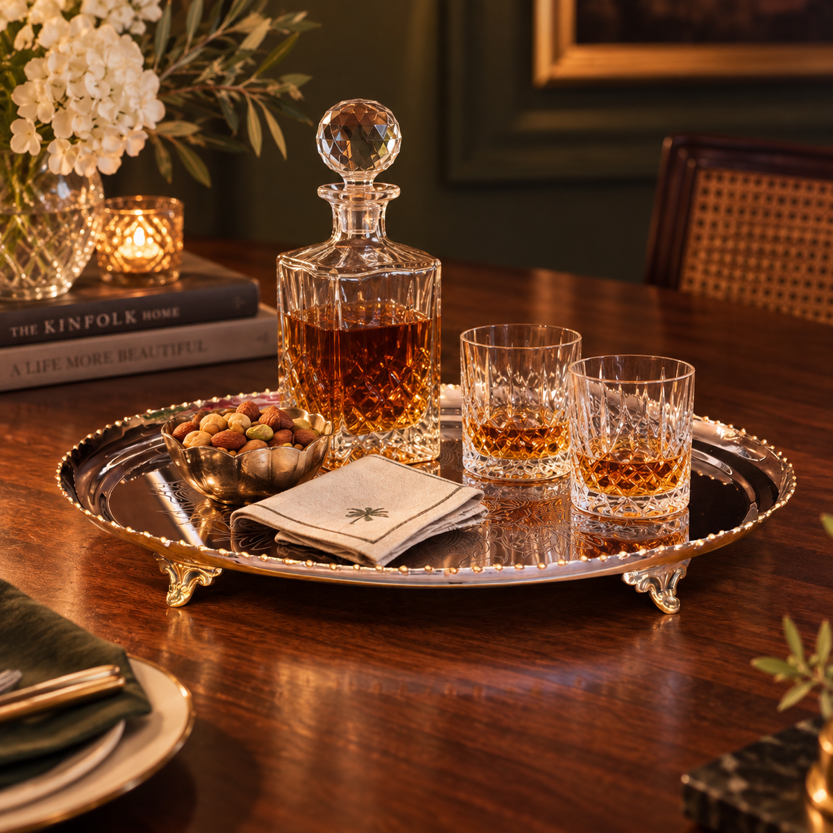 Elegant silver serving tray styled with a crystal whiskey decanter, glasses and nuts on a luxurious wooden dining table.
