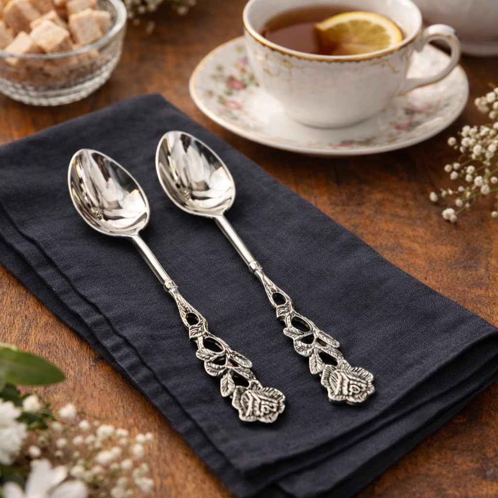 Pair of silver tea spoons with intricate rose floral handles resting on a charcoal grey linen napkin next to a vintage tea cup.