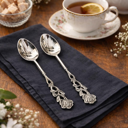 Pair of silver tea spoons with intricate rose floral handles resting on a charcoal grey linen napkin next to a vintage tea cup.