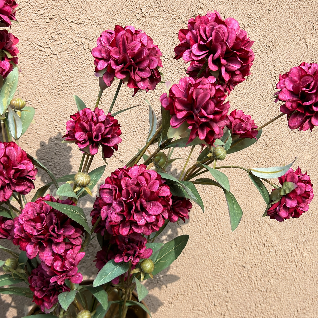 Close-up of artificial burgundy peony flowers with realistic layered petals and green foliage displayed against a textured beige wall, showcasing detailed craftsmanship for luxury home décor.