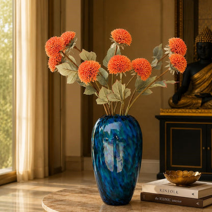 Artificial orange dahlia stems displayed in a blue glass vase on a luxury living room table, creating a warm and elegant home décor arrangement.