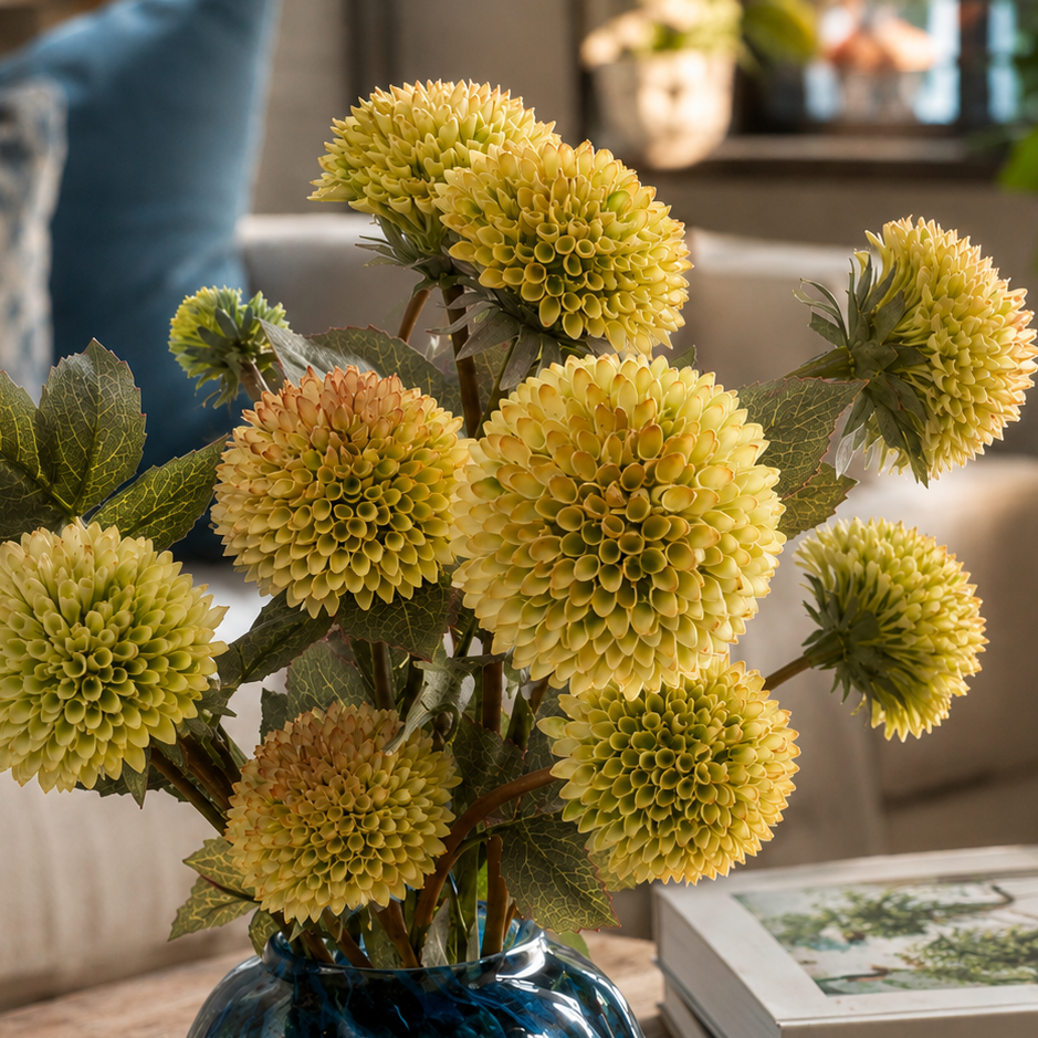 Close-up of realistic artificial white dahlia stems with full layered blooms and natural-looking foliage in a decorative blue glass vase.