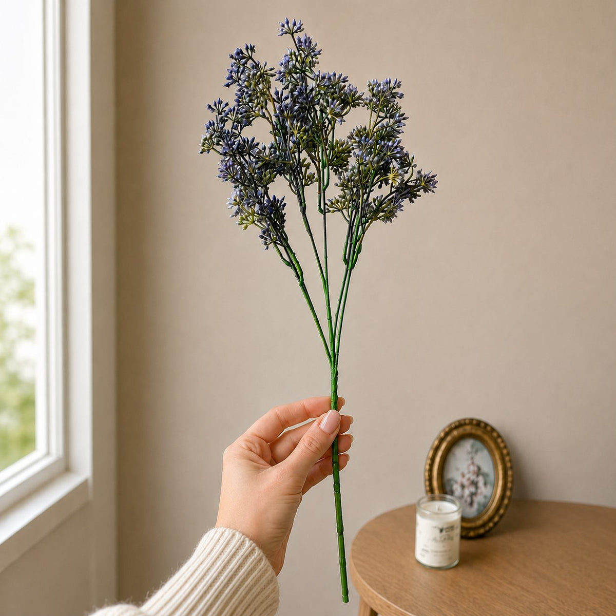 Close-up lifestyle image of a hand holding a single lavender artificial flower stem against a neutral beige wall with soft natural light and minimal home décor in the background.