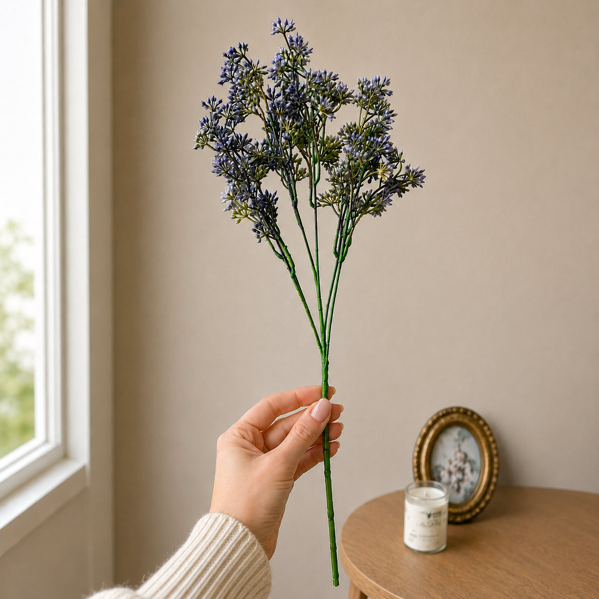 Close-up lifestyle image of a hand holding a single lavender artificial flower stem against a neutral beige wall with soft natural light and minimal home décor in the background.
