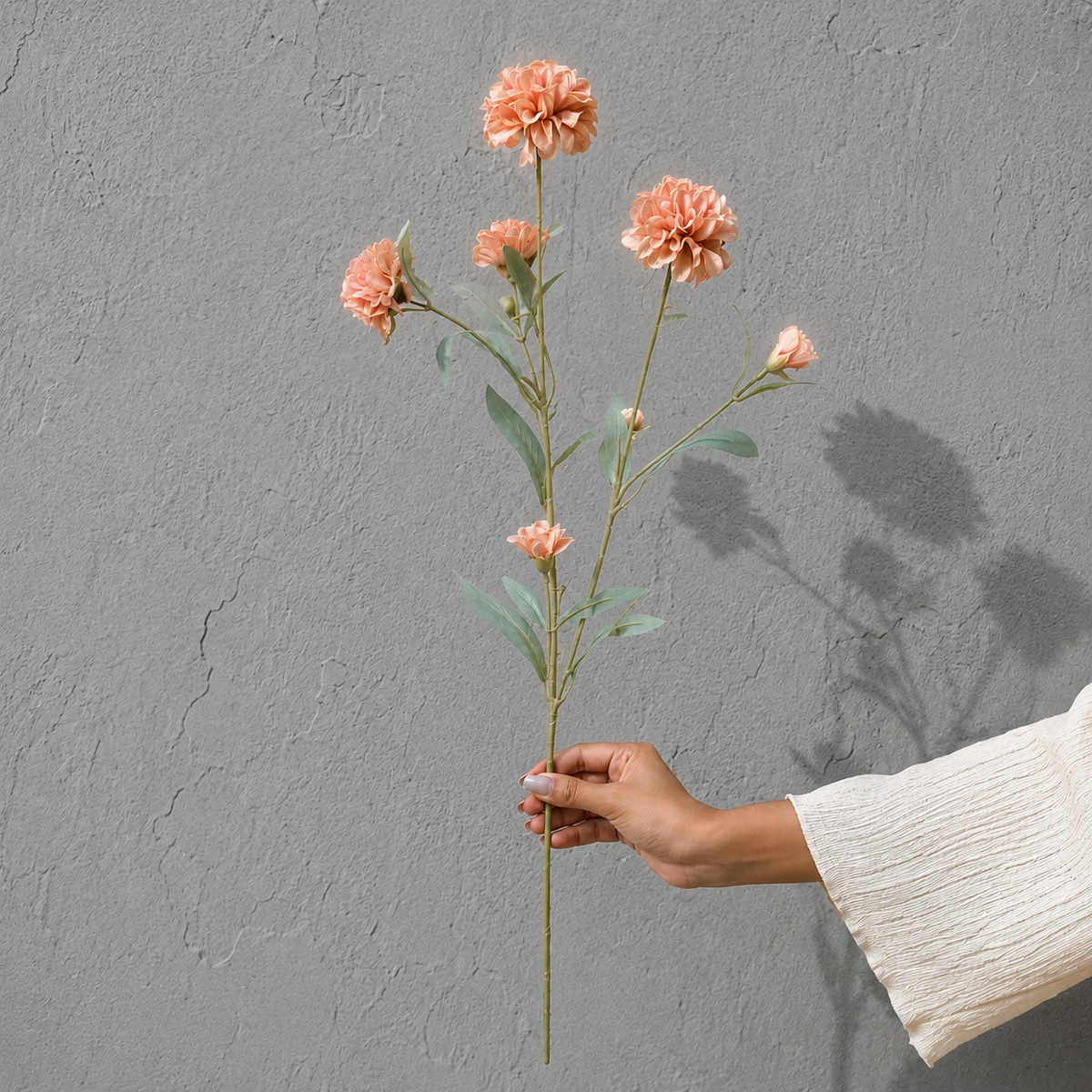 Hand holding a single artificial peach pink flower stem against a textured grey wall, showcasing the size and details of the floral stem in natural light.