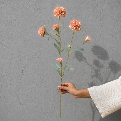 Hand holding a single artificial peach pink flower stem against a textured grey wall, showcasing the size and details of the floral stem in natural light.