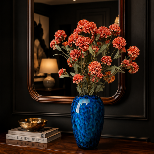 Artificial orange carnation flower stems arranged in a blue glass vase on a wooden console table with a luxury interior setting and decorative mirror.