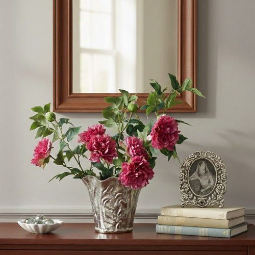 Artificial pink peony flower stems arranged in a decorative silver vase on a wooden console table for elegant home décor.