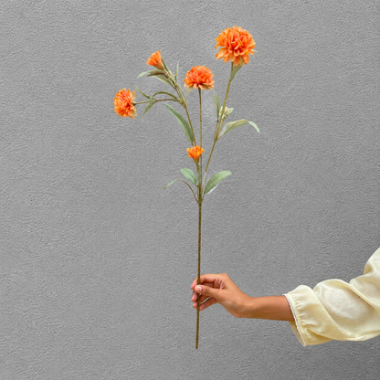 Hand holding a single artificial orange carnation stem with multiple blooms against a textured grey wall, showcasing the flower's size and detailing in natural light.