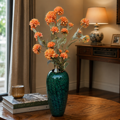 Artificial orange dahlia flower arrangement in a green decorative vase displayed on a wooden coffee table in a modern living room with warm ambient lighting and a clean, elegant background.