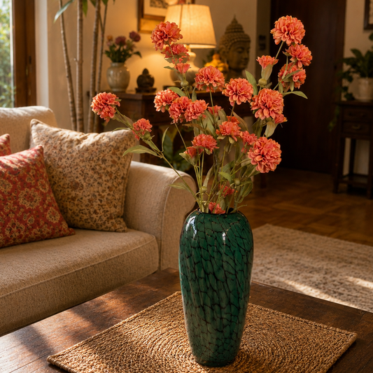 Artificial pink dahlia flower stems arranged in a green glass vase on a coffee table in a cozy living room with a beige sofa and warm natural light.