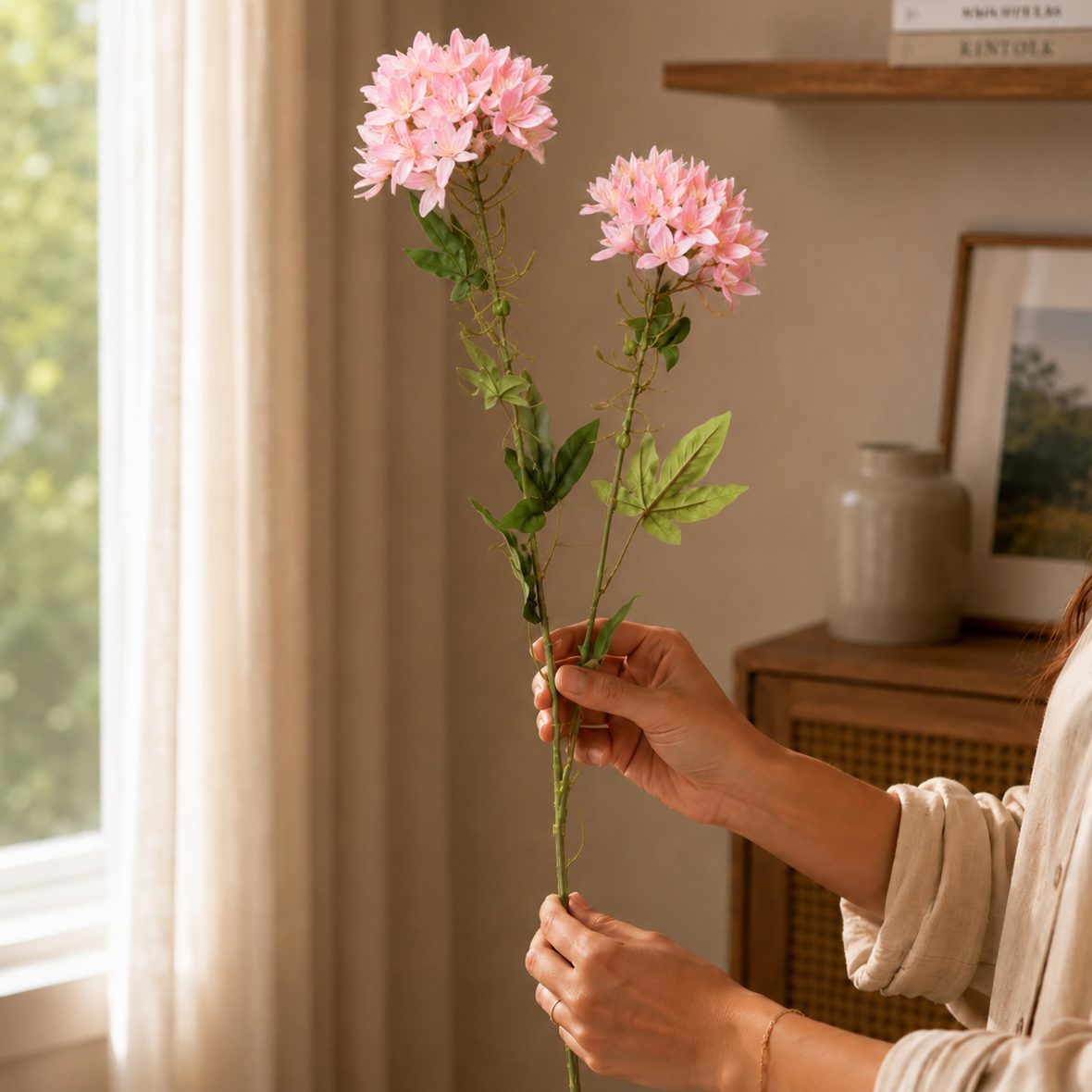 Woman holding pink artificial flower stems in a warm neutral living room setting with soft natural light and elegant home decor background.