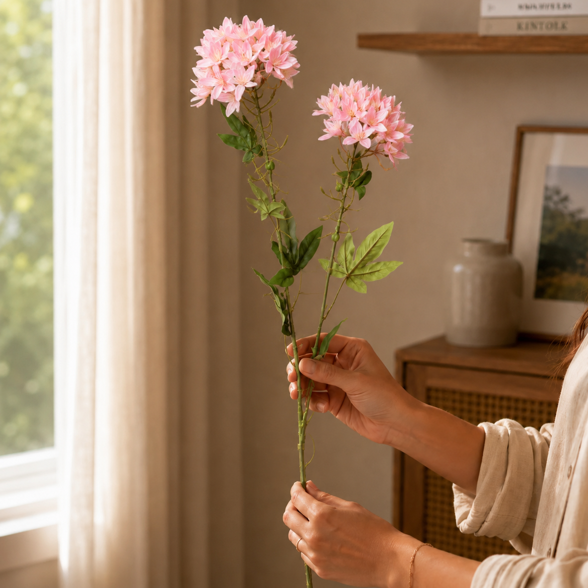 Woman holding pink artificial flower stems in a warm neutral living room setting with soft natural light and elegant home decor background.