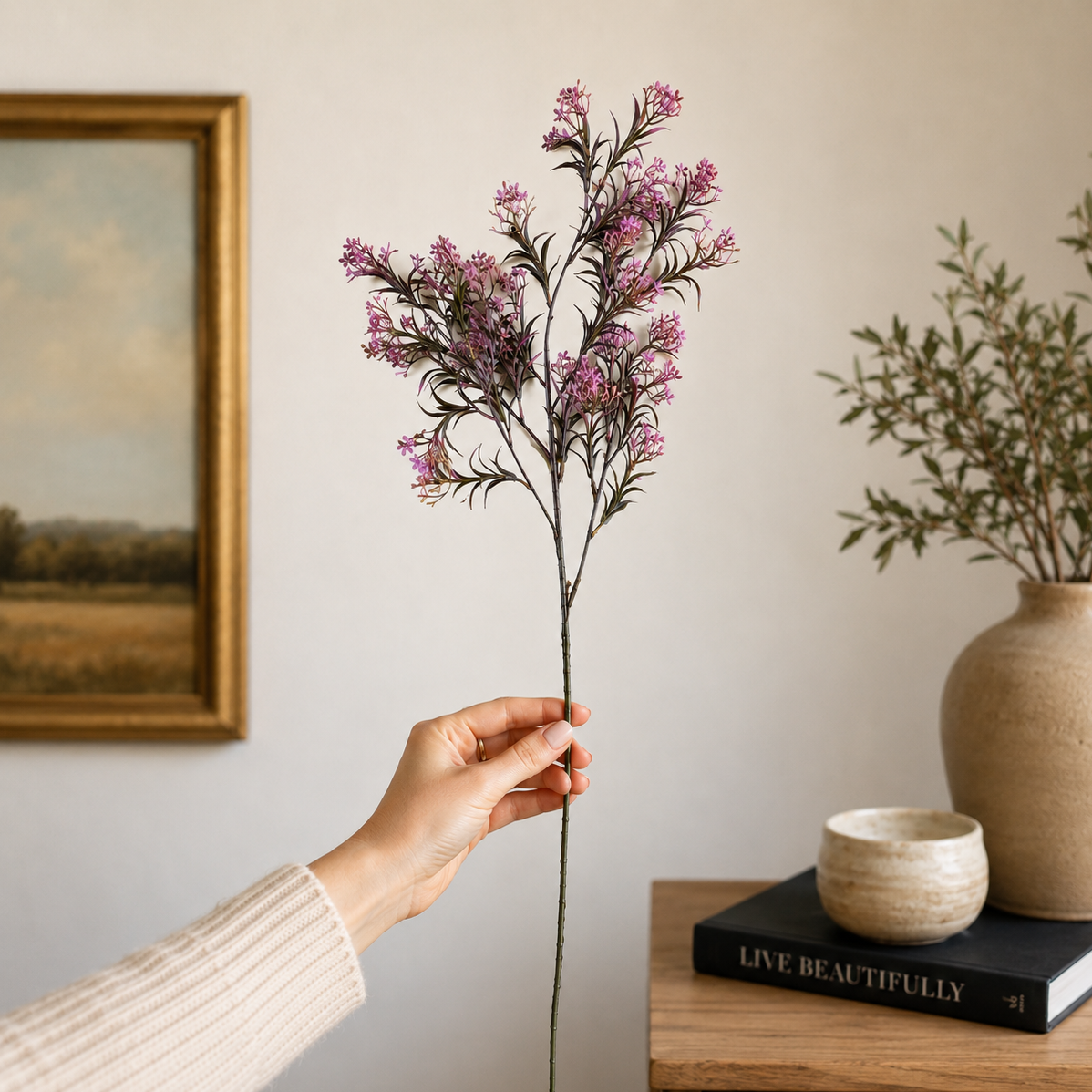 Hand holding a single artificial purple floral stem against a neutral wall in a modern home decor setting, showcasing the size and realistic detailing of the flower stem