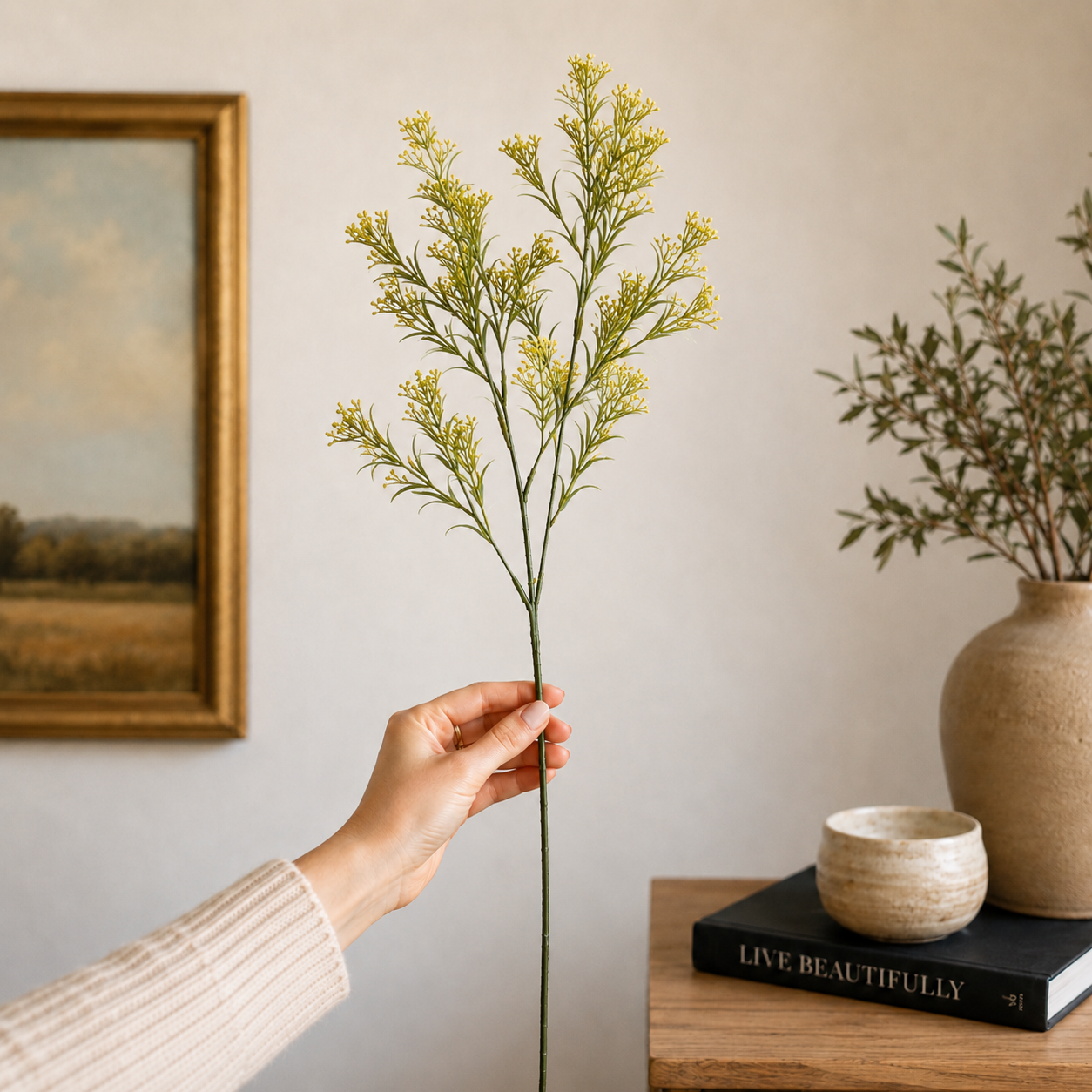 Hand holding a single artificial yellow wildflower stem in a neutral home decor setting with a wooden console table, ceramic vase, and framed wall art in the backgrou