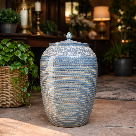 An artisanal blue ceramic floor jar with intricate floral and ribbed textures, styled in a warm, high-end living room next to a wicker planter.