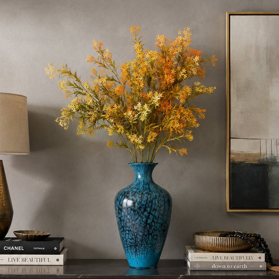 Artificial autumn floral arrangement with orange and cream blossoms displayed in a textured blue glass vase on a modern console table against a grey wall, styled with decorative books, a lamp, and contemporary wall art.