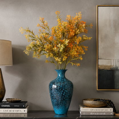 Artificial autumn floral arrangement with orange and cream blossoms displayed in a textured blue glass vase on a modern console table against a grey wall, styled with decorative books, a lamp, and contemporary wall art.